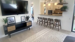 Una sala moderna con un televisor de pantalla plana, una mesa consola con adornos, un tapete estampado y una zona de cocina con taburetes de bar.