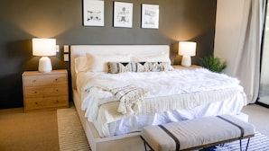 Un dormitorio con una cama grande, dos mesitas de noche con lámparas, un banco y tres cuadros enmarcados en la pared.
