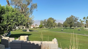 Un campo de golf con vistas despejadas al green, un área de patio con mobiliario de mimbre y una cadena montañosa al fondo.