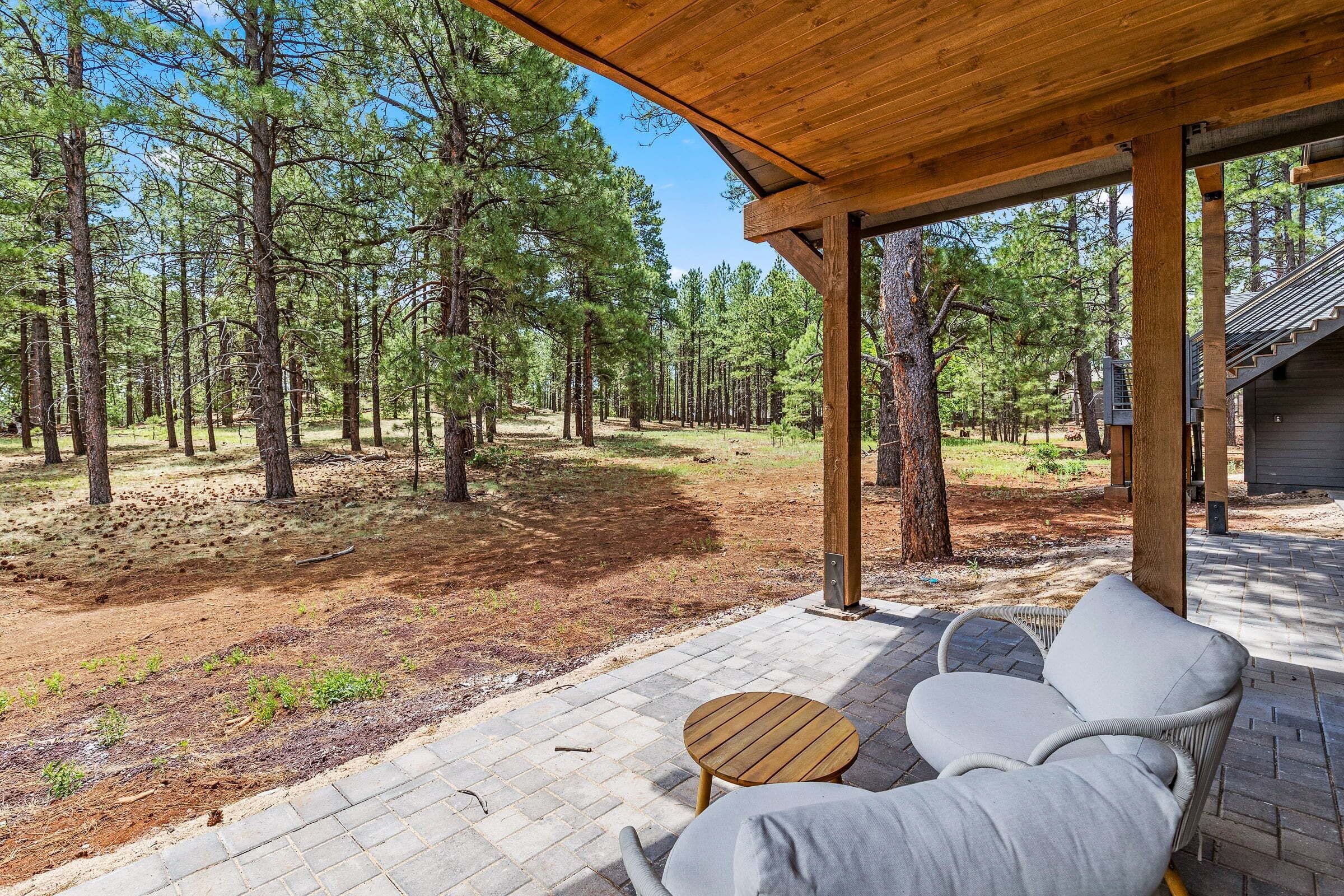 A covered outdoor seating area with a wooden table and two chairs, surrounded by a forested landscape.