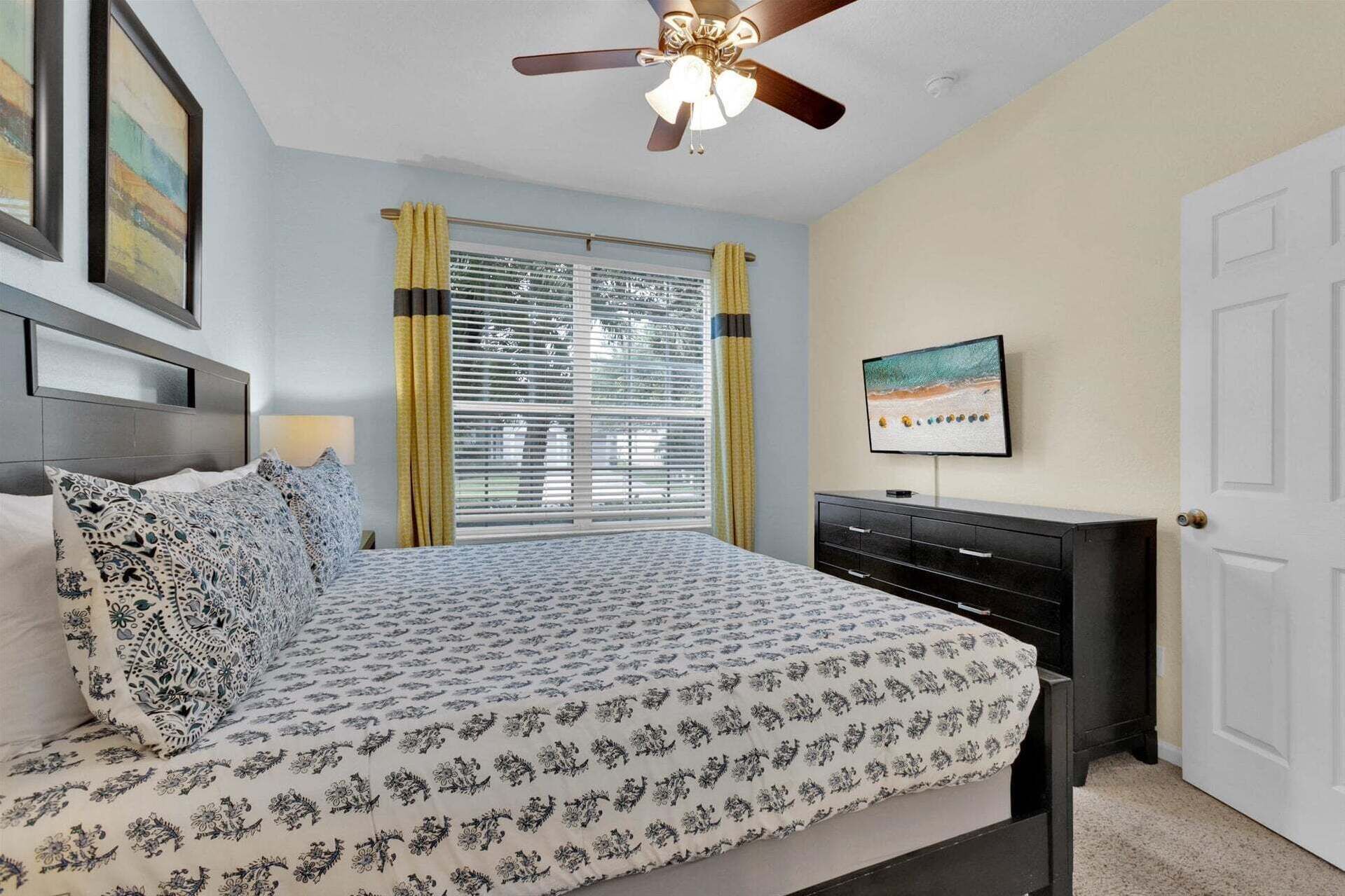 A bedroom with a bed, a ceiling fan, a window with blinds, a TV on the wall, and a door.