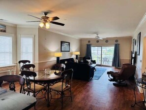 A spacious living room with a dining area, featuring a ceiling fan, a sofa set, and a large window with curtains. - Magnolia Pointe Luster Leaf 4894-301 (Myrtle Beach)