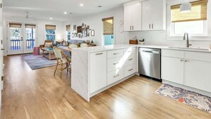 A modern kitchen with white cabinets, a stainless steel dishwasher, and a dining area with wicker chairs. - Little Wing (Santa Rosa Beach)