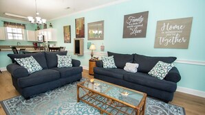 A living room with two dark sofas, a glass coffee table, and patterned rugs. There are wall art pieces and a kitchen in the background. - Magnolia Place 4789-103 (Myrtle Beach)