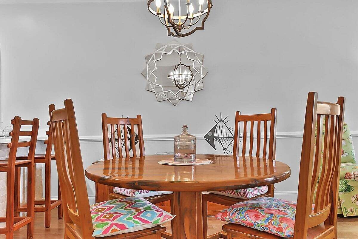 A round wooden dining table with six chairs, each with colorful cushions. Above the table, there are two wall-mounted light fixtures and a geometric wall art piece.