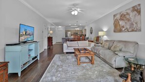 A living room with a sofa, coffee table, and TV. There is a dining area in the background. - Magnolia Pointe Luster Leaf 4825-102 (Myrtle Beach)
