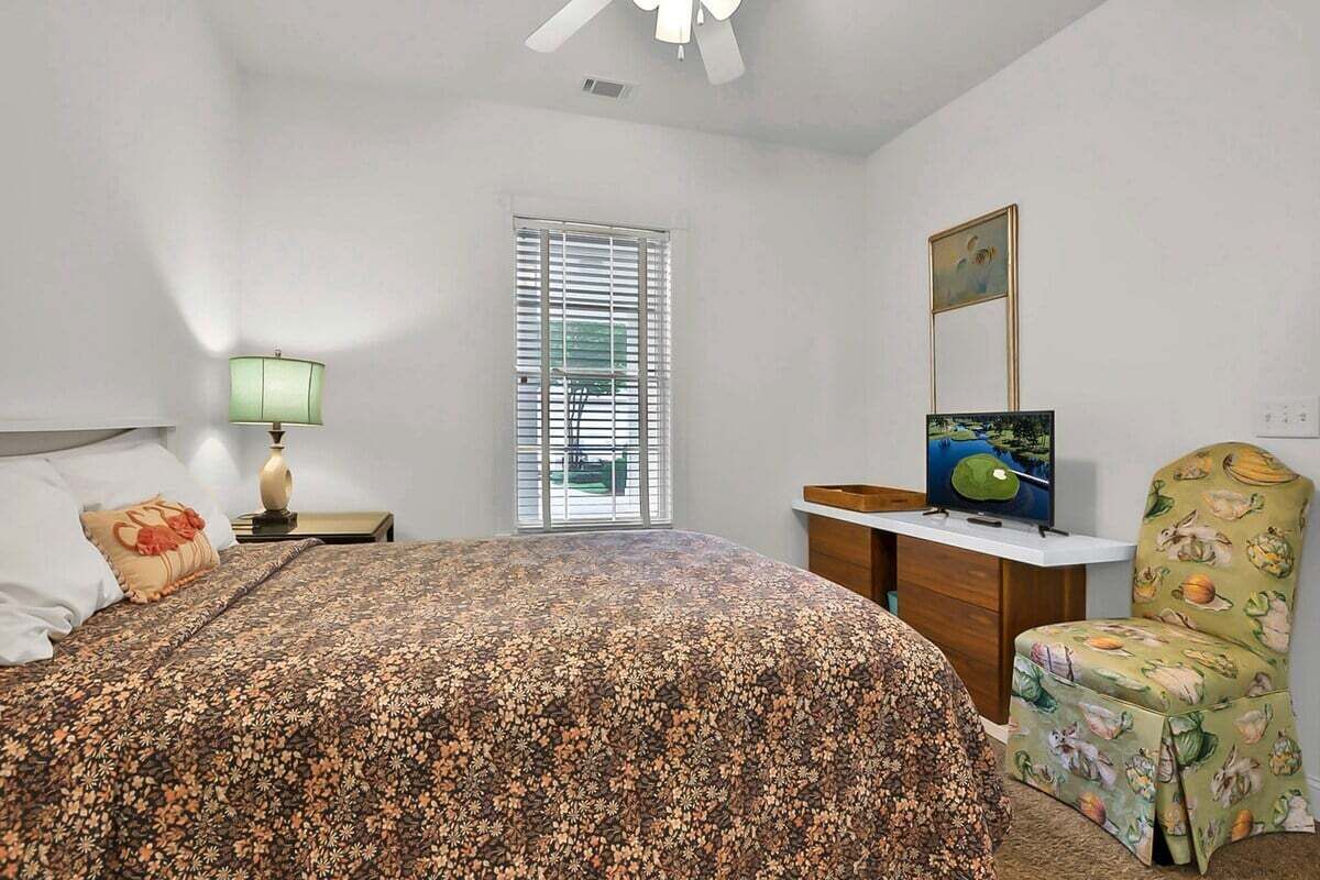 A bedroom with a bed, a desk, a chair, a television, and a window with blinds.