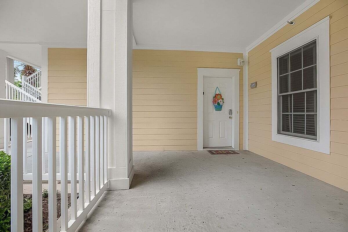 A covered porch with a white railing, a door with a decorative hanging, and a window with blinds.