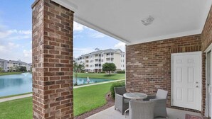 A covered patio with wicker furniture, a brick wall, and a view of a swimming pool and apartment buildings. - Magnolia Pointe Luster Leaf 4825-102 (Myrtle Beach)