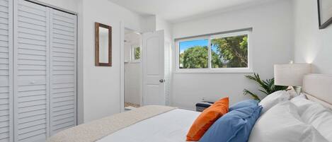 Ein Schlafzimmer mit Bett, Kissen, einem Fenster, einer Tür und einem Nachttisch.