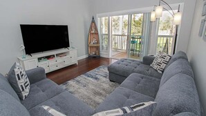 Sala de estar con un sofá gris, televisor de pantalla plana, estante con forma de barco de madera y puerta corrediza de vidrio que da a un patio exterior.
