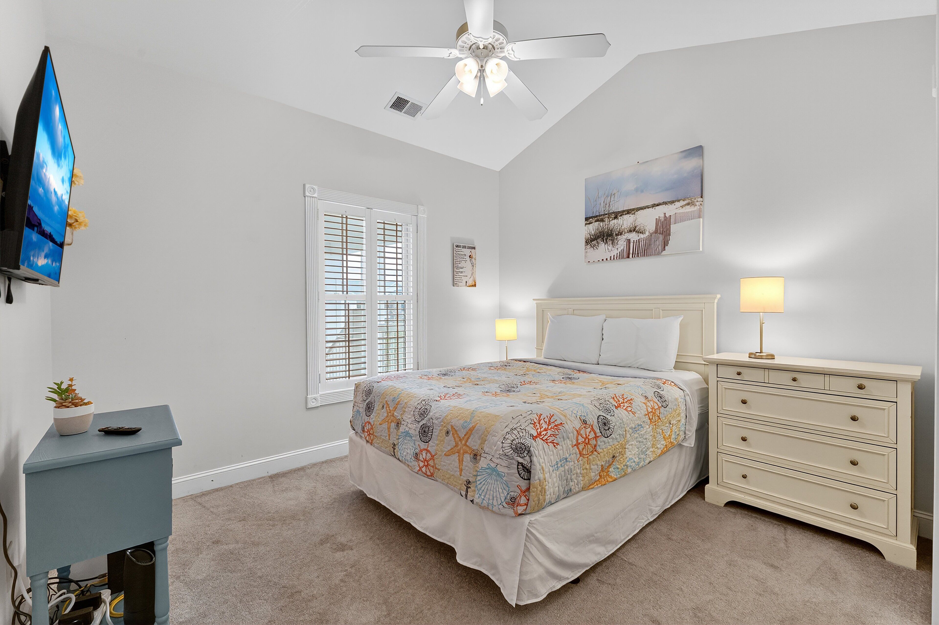 A bedroom with a bed, bedside tables, a ceiling fan, a window with blinds, and a wall-mounted TV.