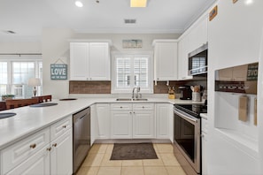 A modern kitchen with white cabinetry, a central island, a built-in oven, and a window with blinds. - Magnolia Pointe 4887-305 (Myrtle Beach)
