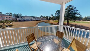 A covered outdoor area with a glass table and wooden chairs, overlooking a body of water and a golf course. - Magnolia North 4839-204 (Myrtle Beach)