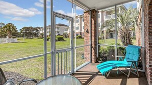 Een balkon met een glazen deur, een blauwe loungestoel en uitzicht op een grasveld met huizen op de achtergrond.