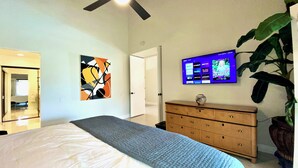 A modern bedroom with a large bed, a dresser, a flat-screen TV mounted on the wall, and a door leading to another room. - Modern Life (Rancho Mirage)