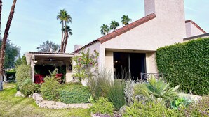 A house with a covered patio, a garden with various plants, and a clear sky. - Modern Life (Rancho Mirage)