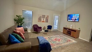 A living room with a brown leather couch, a blue blanket, a patterned rug, a wooden TV stand, and a TV displaying a show. - Modern&beautiful,pool,a+ Location,longstaydiscount (Sherman)