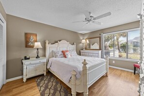 A bedroom with a white bed, a ceiling fan, a window with a view, and a nightstand with a lamp. - Windsor Court 11C (Myrtle Beach)