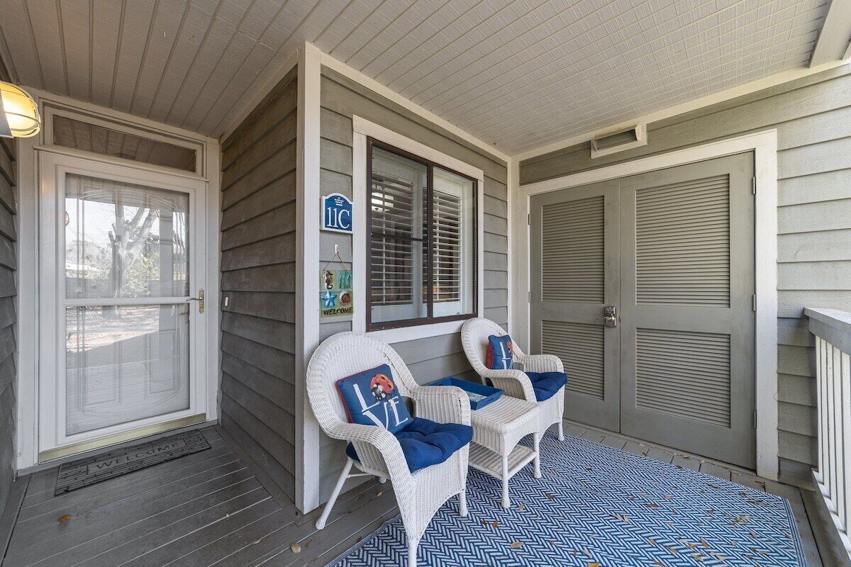 A porch with two wicker chairs, a welcome mat, and a blue patterned rug.