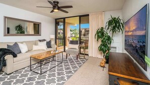 A modern living room with a sofa, coffee table, and a large TV. There is a ceiling fan, a plant, and a view of the outdoors through a sliding door. - Luxury Maui Condo Ocean Views Resort Perks (Kihei)