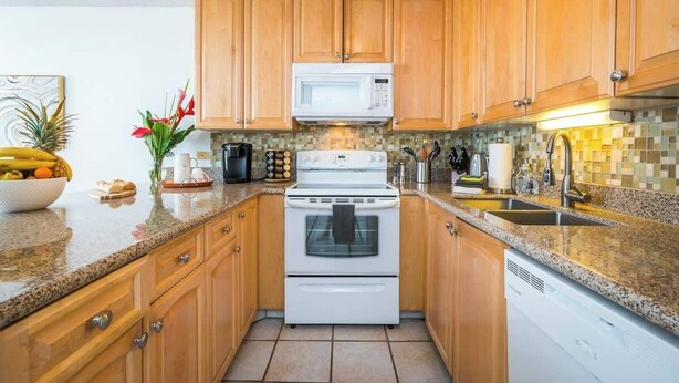 A kitchen with wooden cabinets, a white microwave, a stove, and a dishwasher. - Luxury Maui Condo Ocean Views Resort Perks (Kihei)