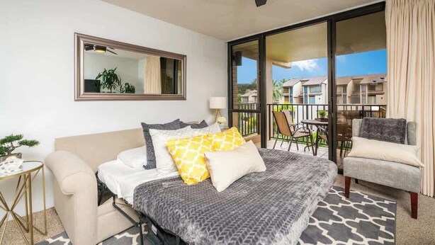A bedroom with a bed, a chair, a small table, a ceiling fan, and a sliding glass door leading to an outdoor area with a view of other buildings. - Luxury Maui Condo Ocean Views Resort Perks (Kihei)