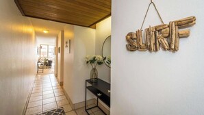 A hallway with a wooden ceiling, a black console table with a vase of white flowers, and a wall-mounted wooden sign spelling "SURF". - Luxury Maui Condo Ocean Views Resort Perks (Kihei)
