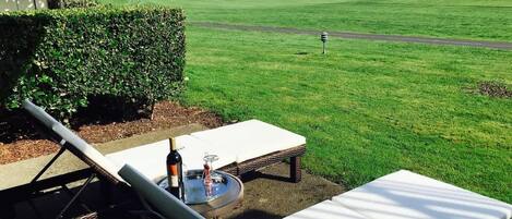 A patio with a lounge chair, a small table with a bottle and glass, and a view of a golf course.