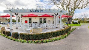 Un espace extérieur aménagé avec des chaises blanches et des parasols rouges, portant l’inscription « MARCHÉ », entouré de haies et agrémenté d’un foyer.