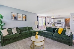 A living room with a green sectional sofa, a round coffee table, and a kitchen in the background.
