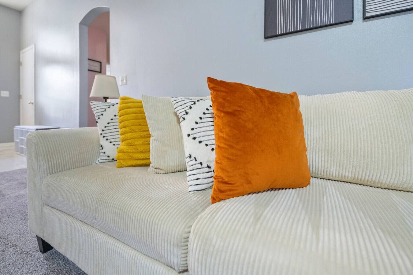 Un canapé de couleur claire, agrémenté de coussins orange à rayures et unis, une lampe et un tableau encadré au mur.