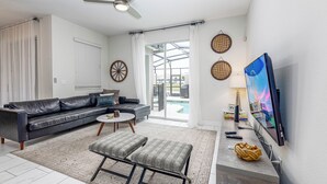 A modern living room with a sofa, ottoman, TV, and a view of a pool. - Tvpm-4885rc/sl (Kissimmee)