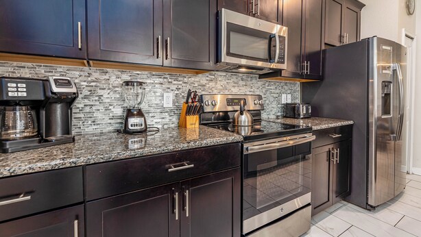 A modern kitchen with dark wood cabinets, stainless steel appliances, a granite countertop, and a tiled backsplash. - Tvpm-4885rc/sl (Kissimmee)