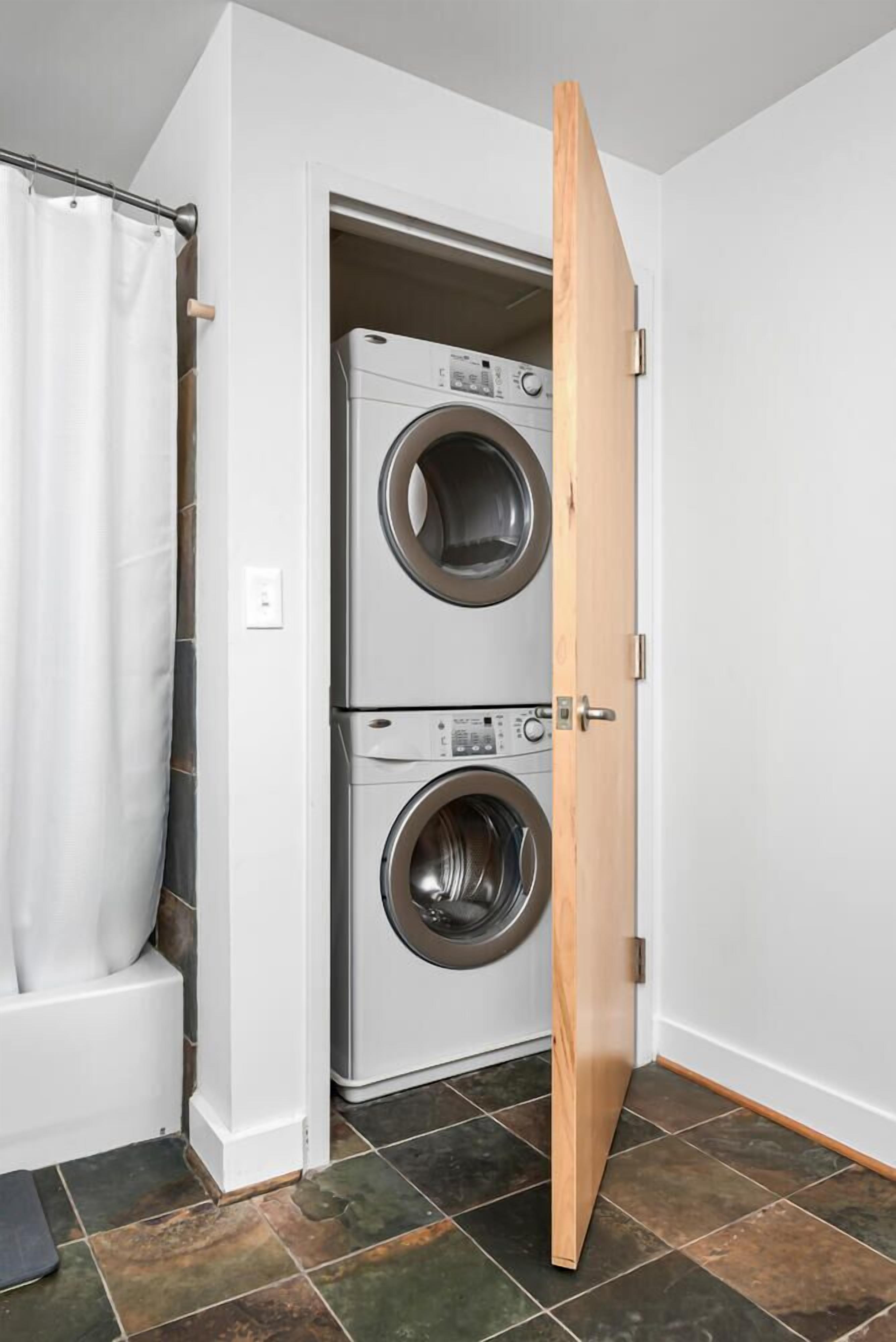 A laundry room with two stacked front-loading washing machines, a white shower curtain, and a tiled floor.