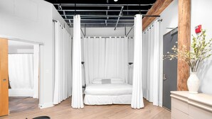 Une chambre à coucher avec un lit, des rideaux blancs, un plancher en bois, une petite table et une commode.