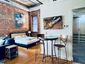 A modern hotel room with a brick wall, a bed, a small table with chairs, and a painting. - Kenny (Nashville)