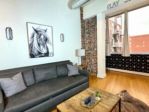 Ein Wohnzimmer mit einem dunkelgrauen Sofa, einem Couchtisch aus Holz mit Tablett, einem an der Wand hängenden Bild von einem Pferd, einer Ziegelwand und Blick auf Gebäude durch eine offene Tür.