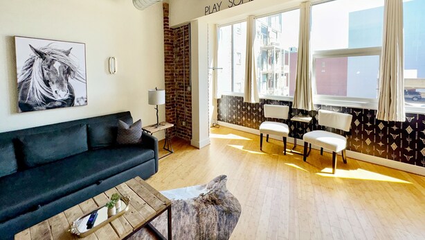 Un salon moderne avec un canapé foncé, une table basse et une cheminée. Une grande fenêtre, ornée de rideaux, offre une vue sur les bâtiments environnants.