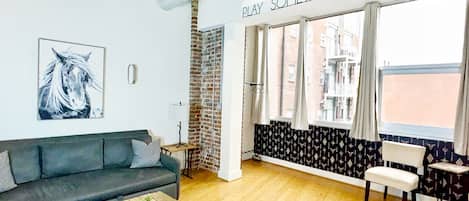 A modern living room with a grey sofa, a wooden coffee table, and a rug. There is a large window with patterned curtains and a sign above that reads "PLAY SOMETHING COUNTRY".