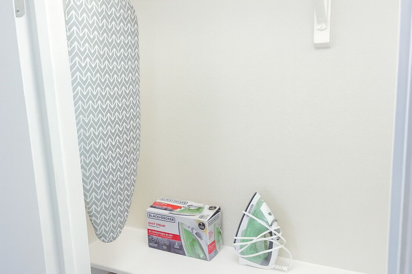 Un coin de pièce avec un mur blanc, un rideau de douche à motifs chevrons gris et blancs, une boîte contenant un fer à vapeur compact Black+Decker Easy Steam, et un fer à repasser vert et blanc.