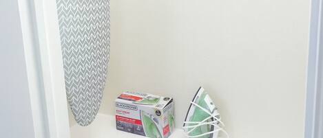 Un coin de pièce avec un mur blanc, un rideau de douche à motifs chevrons gris et blancs, une boîte contenant un fer à vapeur compact Black+Decker Easy Steam, et un fer à repasser vert et blanc.