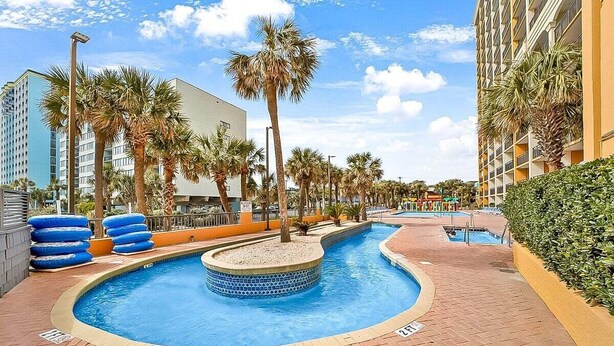 A swimming pool with palm trees and a brick walkway, surrounded by buildings. - Caravelle 706 (Myrtle Beach)