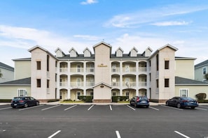 Ein mehrstöckiges Hotel mit zentralem Eingang, zahlreichen Balkonen und einem Parkplatz mit mehreren Autos.
