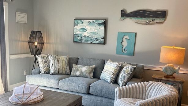 A living room with a grey sofa, a wooden coffee table, a patterned rug, and a decorative fish wall art piece. - Captains Quarters Bright 2BR Pet-friendly Beach Condo w/ Coastal Decor & Relaxed Vibes (Myrtle Beach)