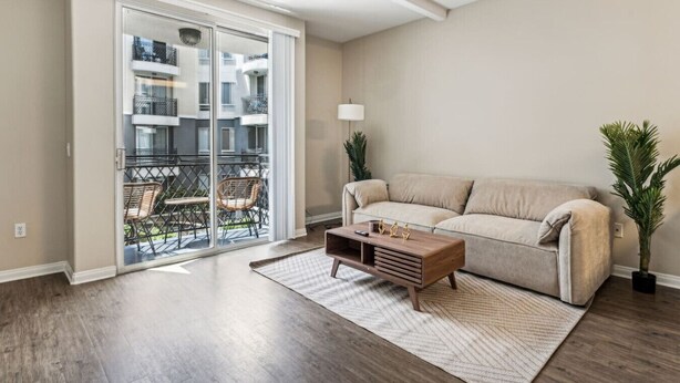 Ein Wohnzimmer mit einer Schiebetür zum Balkon, einem beigen Sofa, einem Holztisch und zwei Blumentöpfen.