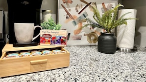Eine Küchenarbeitsplatte mit einer Kaffeemaschine, einer Tasse, einer Auswahl an Teebeuteln, einer Topfpflanze und einem gerahmten Kunstwerk.