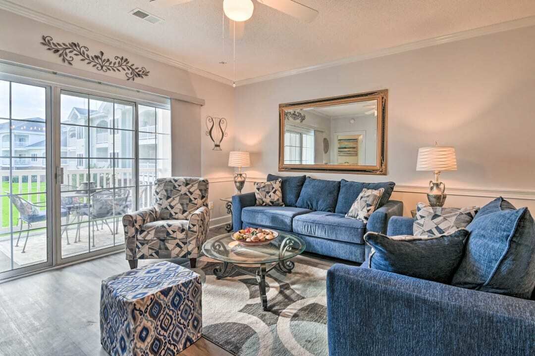 A living room with a blue sofa, patterned armchair, and ottoman. A large mirror and two lamps are on the wall.