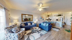 A modern living room with a blue sofa set, a glass coffee table, and a dining area with a white island. - Magnolia North Unit 204 (Myrtle Beach)