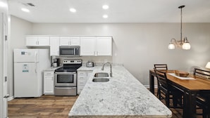 Una cocina con armarios blancos, electrodomésticos de acero inoxidable, encimera de granito y un comedor con sillas y una mesa de madera.